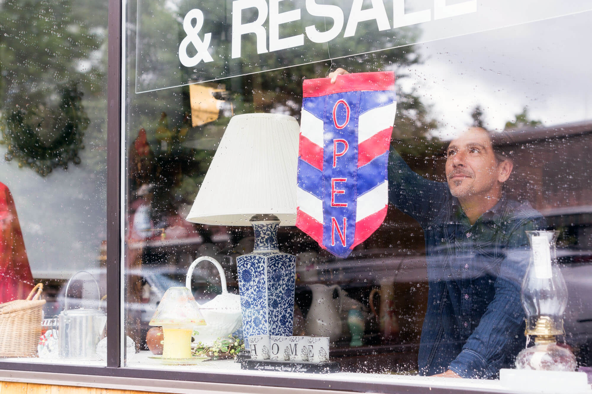 Shop owner putting an "Opened" sign in window while it rains outside.